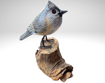 May include: A hand-carved wooden bird sculpture with a gray and tan body, perched on a small, textured wooden branch. The bird has a distinctive crest, black beak, and red eyes. The sculpture is set against a white background.