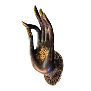 以下が含まれることがあります： 手のひらに金色の模様が施されたブロンズ製の彫刻。手は指を内側に曲げて、与えるか提供するジェスチャーをしています。彫刻は円形の台座に載せられています。