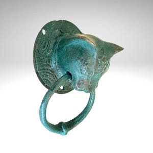May include: A bronze cat-shaped door knocker with a circular handle. The knocker has a weathered, verdigris finish, with a round mounting plate. The cat's head is detailed with textured fur and a pointed ear.