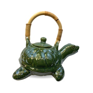 May include: Green ceramic teapot shaped like a turtle with a brown woven handle. The teapot has a detailed design of leaves and flowers.