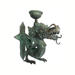May include: A bronze-colored dragon-shaped wall sconce. The dragon has a detailed face with sharp teeth and a long, curling tongue. It holds a small, bowl-shaped cup above its head. The sconce has a decorative, geometric backplate.