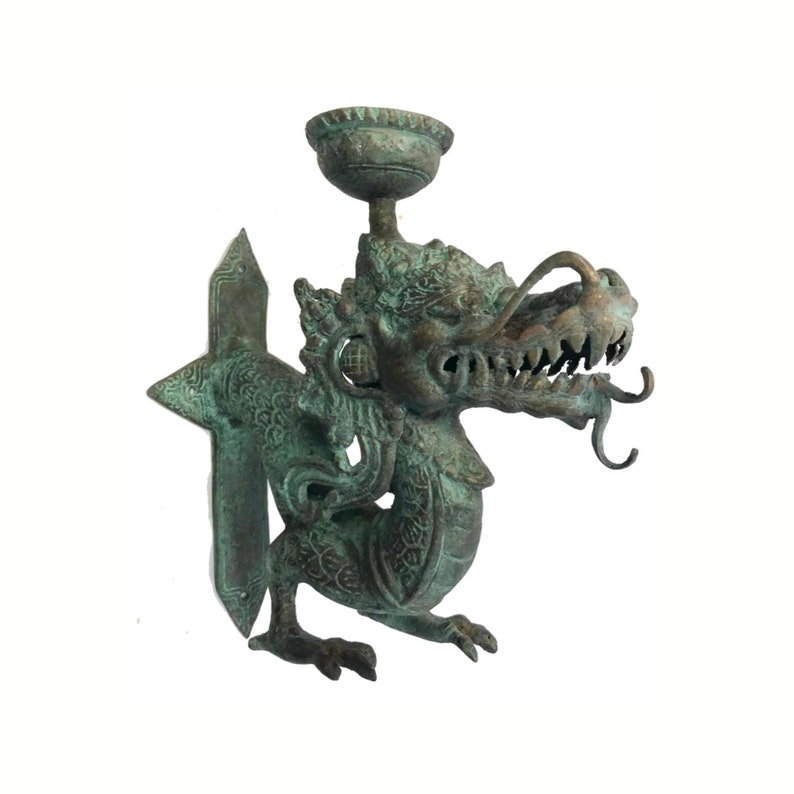 May include: A bronze-colored dragon-shaped wall sconce. The dragon has a detailed face with sharp teeth and a long, curling tongue. It holds a small, bowl-shaped cup above its head. The sconce has a decorative, geometric backplate.