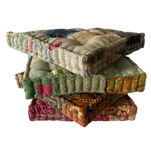 May include: A stack of four square floor cushions. Each cushion is made of patchwork fabric in various colors and patterns, including floral and geometric designs. The cushions have a quilted appearance with a thick, padded border.