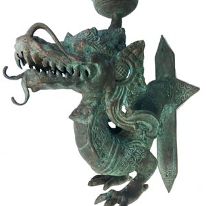 May include: A detailed bronze dragon sculpture with a verdigris finish. The dragon features intricate scales, sharp teeth, and a small cup-like structure on top. The sculpture has a decorative, ornate design.