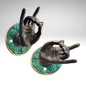 Peut inclure: Deux poignées de porte en bronze en forme de mains faisant un geste. Chaque main est fixée sur une plaque arrière ronde, verte et ornée, avec une bordure dorée. Les mains présentent des détails de jointures et de rides.