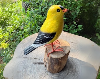 Pode incluir: Escultura de pássaro de madeira pintada à mão. O pássaro tem penas amarelas brilhantes, marcas pretas e brancas nas asas e uma cauda cinza. Ele está em um pequeno toco de madeira com pernas laranja e bico vermelho. Fundo natural.