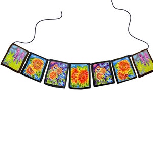 May include: A colorful floral garland with black borders and blue string. The garland features orange, yellow, and red flowers with green leaves and purple accents.