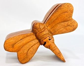Puede incluir: Una caja de rompecabezas de madera con forma de libélula con intrincados tallados. La caja está hecha de madera marrón claro y tiene un acabado natural.