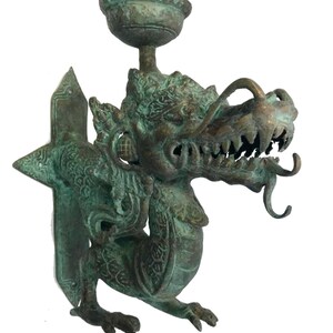 May include: A bronze-colored dragon-shaped wall sconce. The dragon has detailed scales, a fierce expression, and a small bowl at the top. The sconce is designed to hold a candle or small light source, adding a touch of fantasy to any space.
