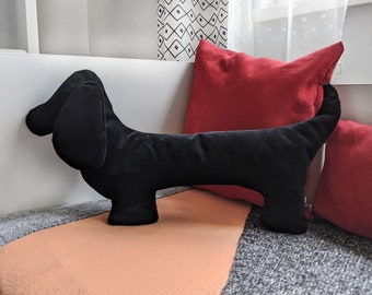 May include: A black plush dachshund dog toy with a long body and short legs. The toy is lying on a gray and orange blanket with a red pillow behind it.