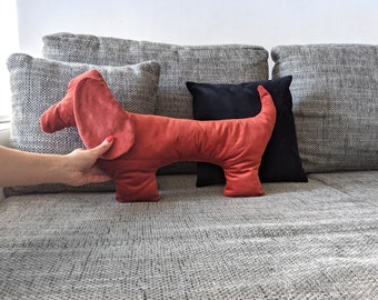 May include: A red dachshund-shaped pillow on a gray couch. The pillow is made of a soft, plush fabric and has a realistic dachshund design.