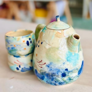 May include: A ceramic teapot and two stacked ceramic cups featuring a cream-colored base with abstract hand-painted patterns in shades of blue, green, and orange. The teapot has a lid with a small handle and a spout, while the cups are cylindrical.