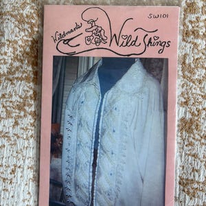 May include: A pink packet featuring the text "Wildman's Wild Things" and a drawing of a person wearing a hat. The packet contains a photograph of a white jacket with embroidery and a blue zipper.