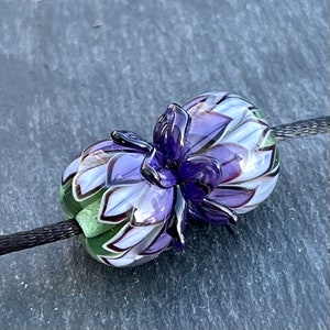 May include: A handcrafted glass bead with a floral design. The bead features a deep purple flower with white and lavender petals. The bead is strung on a black cord, showcasing the intricate details and colors.