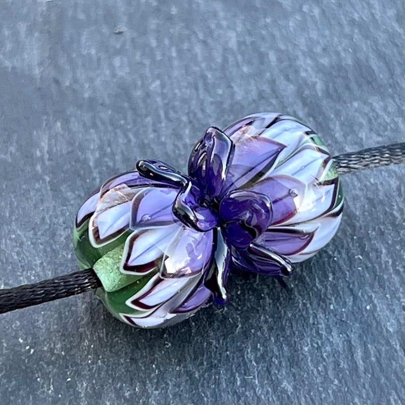 May include: A handcrafted glass bead with a floral design. The bead features a deep purple flower with white and lavender petals. The bead is strung on a black cord, showcasing the intricate details and colors.