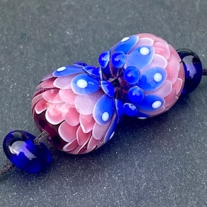 Op de afbeelding: Twee glazen kralen met een roze en blauwe kleurstelling. De kralen zijn versierd met witte stippen en een bloemvormig patroon. De kralen zijn geregen aan een zwart koord.