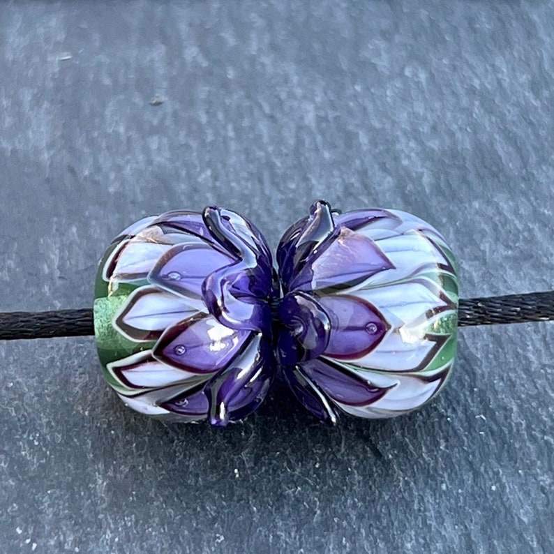 May include: Two glass beads with a floral design. The beads are round, with a purple, white, and green color scheme. The beads are strung on a black cord. The beads have a flower-like design.