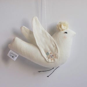 Può includere: Ornamento a forma di uccello in tessuto bianco fatto a mano con un piccolo fiore sulla testa e accenti di perline su un'ala. L'uccello ha zampe di filo nero e un'etichetta con la scritta "JUMATA MADE".