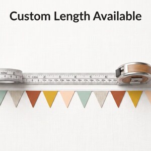 Puede incluir: Una guirnalda de banderines triangulares en tonos apagados de verde salvia, beige, naranja &oacute;xido y amarillo mostaza. Una cinta m&eacute;trica se sit&uacute;a sobre los banderines, mostrando cent&iacute;metros, con el texto Custom Length Available.