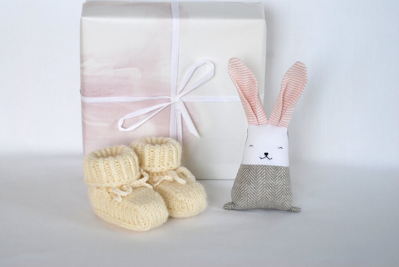 May include: A gift set for a new baby, featuring a white gift box with a pink and white ribbon, a pair of knitted cream booties, and a gray and white bunny toy with pink ears.