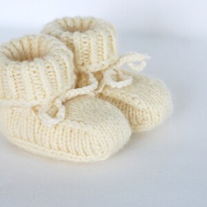May include: A pair of knitted baby booties in a cream color with a tied bow on each bootie.