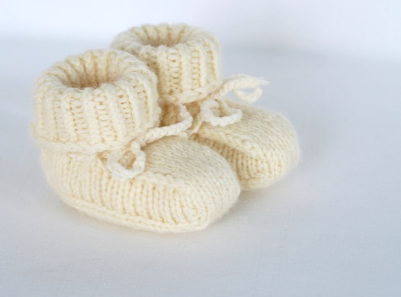 May include: A pair of knitted baby booties in a cream color with a tied bow on each bootie.