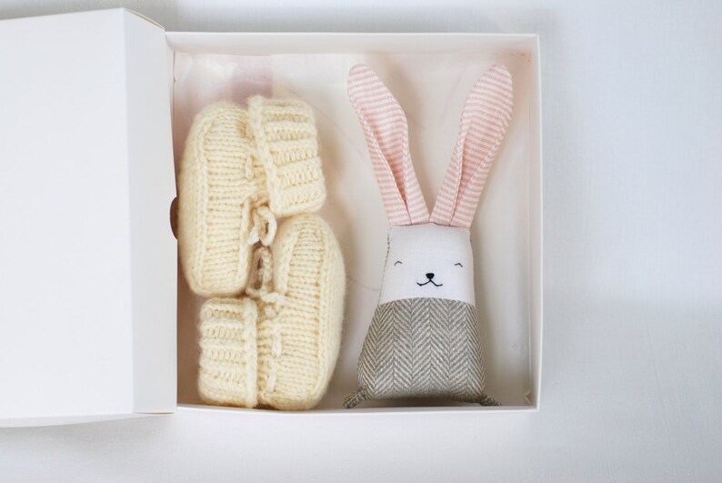 May include: A white gift box with a pink and white striped bunny toy and a pair of cream knitted baby booties.