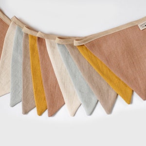 May include: A bunting banner with ten triangular flags in shades of beige, light brown, gray, and yellow. The flags are made of a linen-like fabric and are strung together with a beige ribbon.