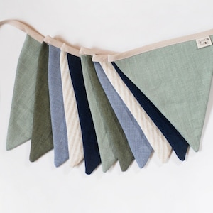 Può includere: Una ghirlanda di bandierine in tessuto con triangoli nei toni del verde salvia, oliva, blu navy e azzurro, alternate a bandierine a righe color crema. Le bandierine sono fissate su un nastro beige con una piccola etichetta JUMAT MADE.