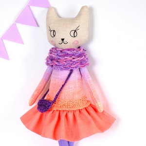 Può includere: Una bambola di gatto fatta a mano con un vestito rosa e viola sfumato, una sciarpa all'uncinetto viola e una borsa all'uncinetto viola. La bambola ha un viso beige con occhi ricamati e un naso rosa.