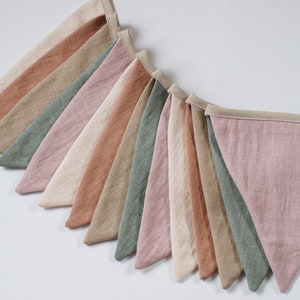 Puede incluir: Una guirnalda de banderines de tela en tonos rosa, verde, beige y marr&oacute;n. Los banderines est&aacute;n hechos de lino y tienen un aspecto sencillo y r&uacute;stico. La guirnalda es perfecta para a&ntilde;adir un toque de fantas&iacute;a a cualquier habitaci&oacute;n.
