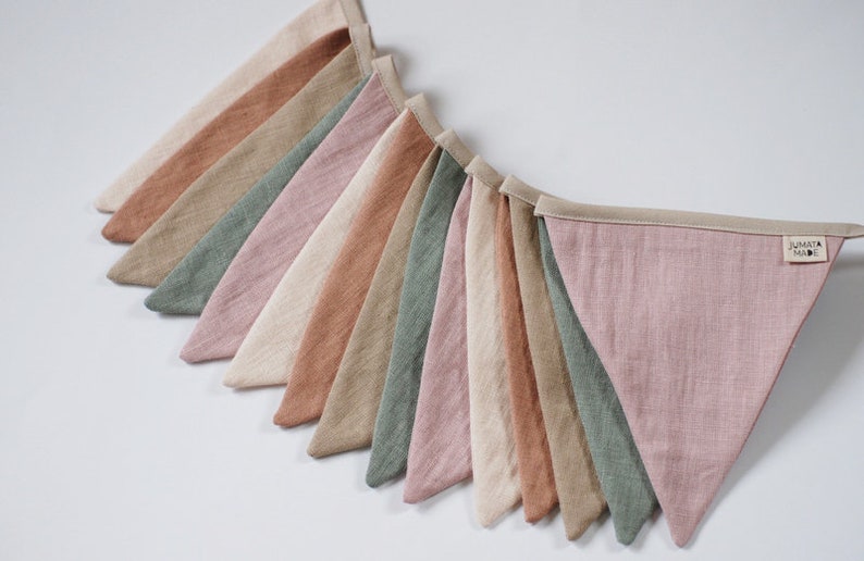 Puede incluir: Una guirnalda de banderines de tela en tonos rosa, verde, beige y marr&oacute;n. Los banderines est&aacute;n hechos de lino y tienen un aspecto sencillo y r&uacute;stico. La guirnalda es perfecta para a&ntilde;adir un toque de fantas&iacute;a a cualquier habitaci&oacute;n.