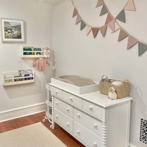 Puede incluir: C&oacute;moda blanca con seis cajones y una mesa para cambiar pa&ntilde;ales en la parte superior. La c&oacute;moda tiene un dise&ntilde;o de patas torneadas decorativas. Una cesta de mimbre con un animal de peluche se encuentra en la mesa para cambiar pa&ntilde;ales. Una hilera de banderas triangulares de colores pastel cuelga de la pared detr&aacute;s de la c&oacute;moda.