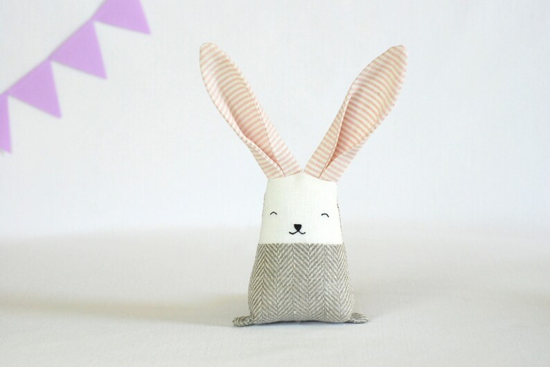 May include: A plush gray and white bunny toy with pink striped ears. The bunny has a simple embroidered face with black eyes and a nose.