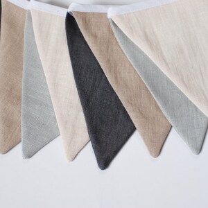 Puede incluir: Una guirnalda decorativa que presenta una serie de banderas de tela triangulares en tonos alternos de gris carb&oacute;n, beige y verde salvia claro. Cada bandera est&aacute; unida a una cinta de cabecera de tela blanca, mostrando una textura de lino visible.