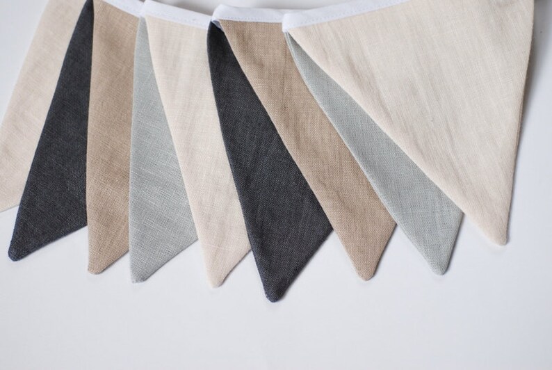 Puede incluir: Una guirnalda decorativa que presenta una serie de banderas de tela triangulares en tonos alternos de gris carb&oacute;n, beige y verde salvia claro. Cada bandera est&aacute; unida a una cinta de cabecera de tela blanca, mostrando una textura de lino visible.