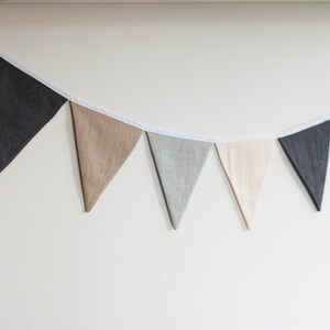 Puede incluir: Una guirnalda de banderines compuesta por una serie de banderas triangulares de tela en tonos alternos de gris oscuro, gris claro, beige y crema. Los banderines est&aacute;n sujetos a una fina cinta de tela blanca que cuelga en arco.