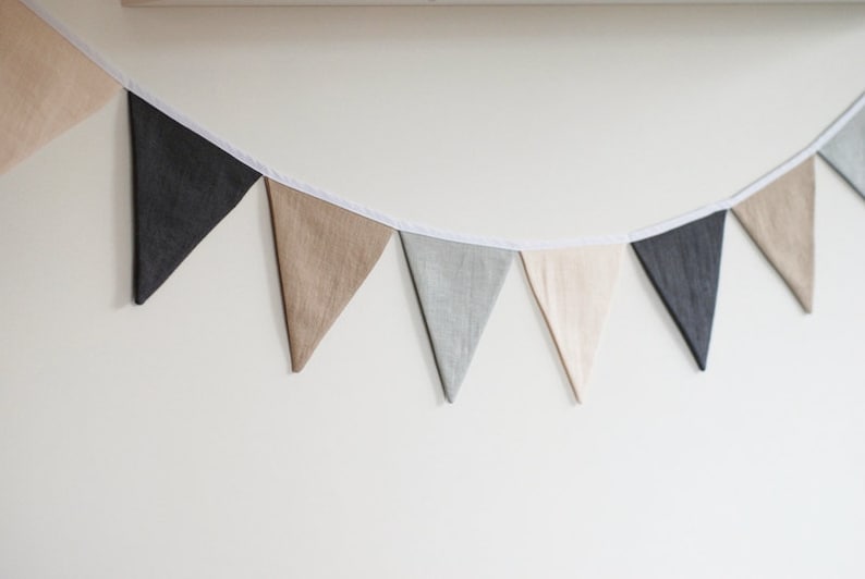 Puede incluir: Una guirnalda de banderines compuesta por una serie de banderas triangulares de tela en tonos alternos de gris oscuro, gris claro, beige y crema. Los banderines est&aacute;n sujetos a una fina cinta de tela blanca que cuelga en arco.