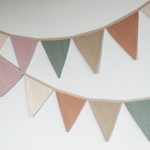 Puede incluir: Una guirnalda de banderines de tela con banderas triangulares en tonos rosa, verde, beige y marr&oacute;n. Las banderas est&aacute;n cosidas juntas con una cuerda blanca.