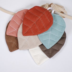 Può includere: Cinque forme di foglie in tessuto di diversi colori, tra cui marrone, beige, rosso, rosa e blu, sono infilate su un cordoncino color crema.