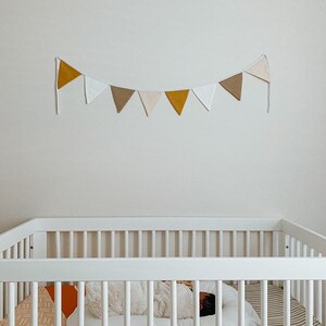 Puede incluir: Una guirnalda de tela con ocho banderines triangulares en tonos alternos de amarillo mostaza, blanco y beige. Los banderines est&aacute;n sujetos a una cuerda blanca y colgados horizontalmente en una pared lisa sobre una cuna blanca.