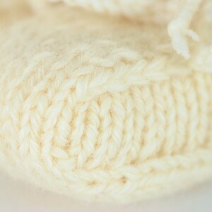 May include: Close-up of a knitted cream-colored fabric with a textured pattern. The fabric is soft and fluffy, with a subtle sheen.