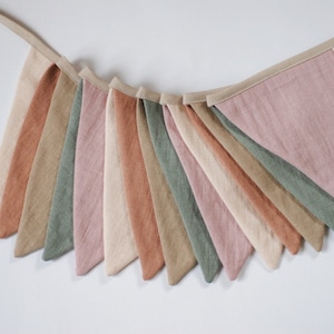 Puede incluir: Una guirnalda de banderines con 12 banderines triangulares en tonos de rosa, verde, beige y marr&oacute;n. Los banderines est&aacute;n hechos de una tela suave y texturizada y est&aacute;n conectados por una cinta beige.