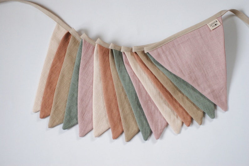Puede incluir: Una guirnalda de banderines con 12 banderines triangulares en tonos de rosa, verde, beige y marr&oacute;n. Los banderines est&aacute;n hechos de una tela suave y texturizada y est&aacute;n conectados por una cinta beige.