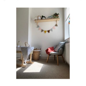 Puede incluir: Una habitaci&oacute;n infantil con un estante de pared de madera y una guirnalda de banderines de tela en tonos rosa, gris, mostaza y marr&oacute;n. Un mois&eacute;s de mimbre con una manta estampada, un cesto de ropa trenzado y un sill&oacute;n gris con una manta roja.