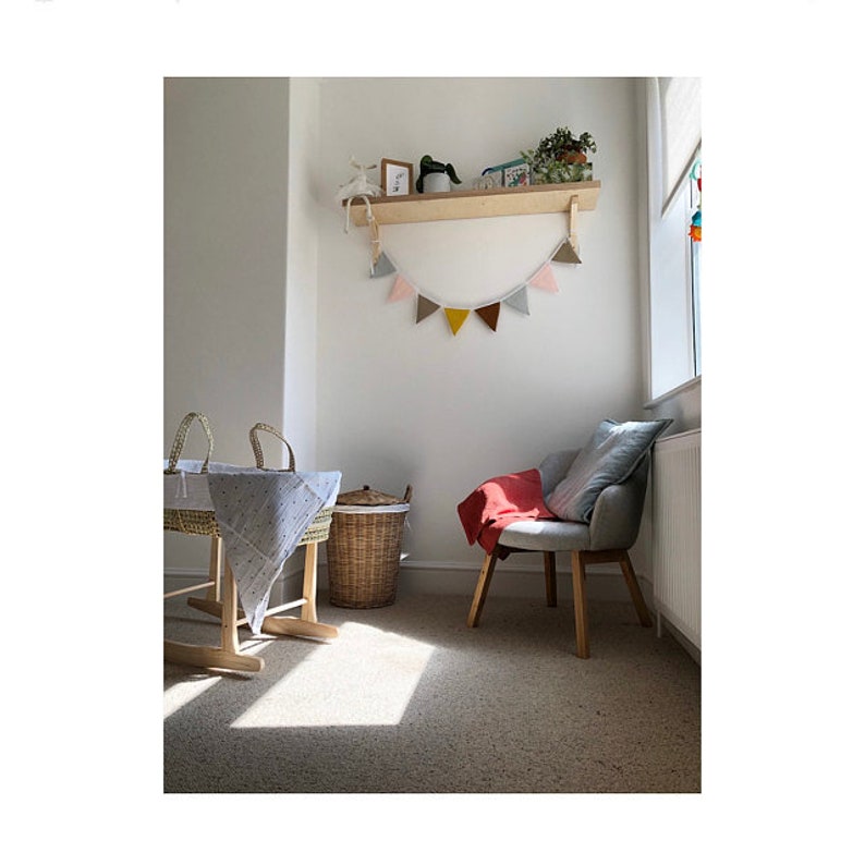 Puede incluir: Una habitaci&oacute;n infantil con un estante de pared de madera y una guirnalda de banderines de tela en tonos rosa, gris, mostaza y marr&oacute;n. Un mois&eacute;s de mimbre con una manta estampada, un cesto de ropa trenzado y un sill&oacute;n gris con una manta roja.