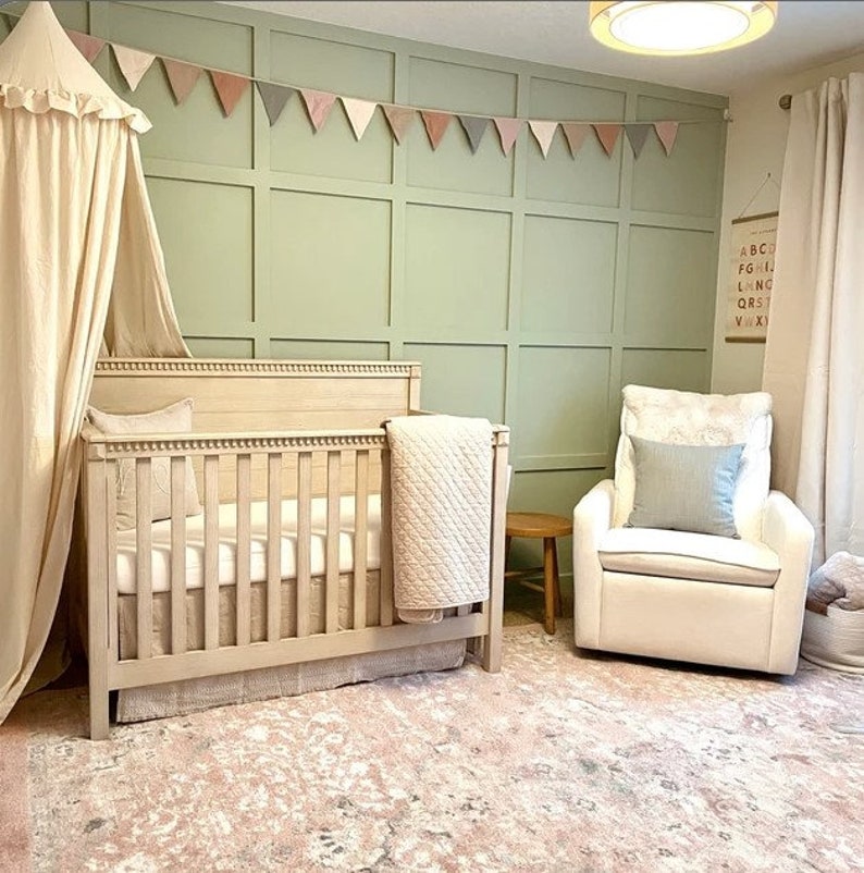 Puede incluir: Una cuna blanca con un dosel blanco y una mecedora blanca en una habitaci&oacute;n de beb&eacute; con una pared verde y una guirnalda de tri&aacute;ngulos rosa y gris.