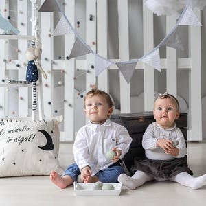 K&ouml;nnte beinhalten: Zwei Babys sitzen auf einem wei&szlig;en Boden. Eines tr&auml;gt ein wei&szlig;es Hemd und Jeans, das andere ein wei&szlig;es Oberteil und einen dunklen Rock. Ein Kissen mit Text und einem Mond liegt auf dem Boden. Ein dekoratives Banner h&auml;ngt dar&uuml;ber.