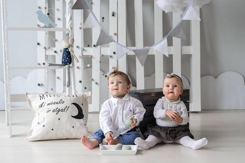 K&ouml;nnte beinhalten: Zwei Babys sitzen auf einem wei&szlig;en Boden. Eines tr&auml;gt ein wei&szlig;es Hemd und Jeans, das andere ein wei&szlig;es Oberteil und einen dunklen Rock. Ein Kissen mit Text und einem Mond liegt auf dem Boden. Ein dekoratives Banner h&auml;ngt dar&uuml;ber.