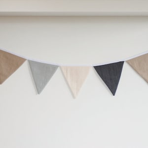 Puede incluir: Una guirnalda de banderines con siete banderas triangulares de tela en tonos alternos de gris carb&oacute;n, beige, gris claro y crema. Las banderas est&aacute;n unidas a una cinta de tela blanca continua, creando un adorno colgante decorativo.